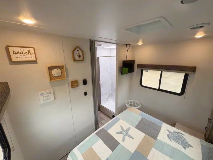 This RV rental offers a cozy bedroom with beach-themed decor, a comfortable bed, TV, and an adjacent shower.. Gulf Stream Conquest 2025