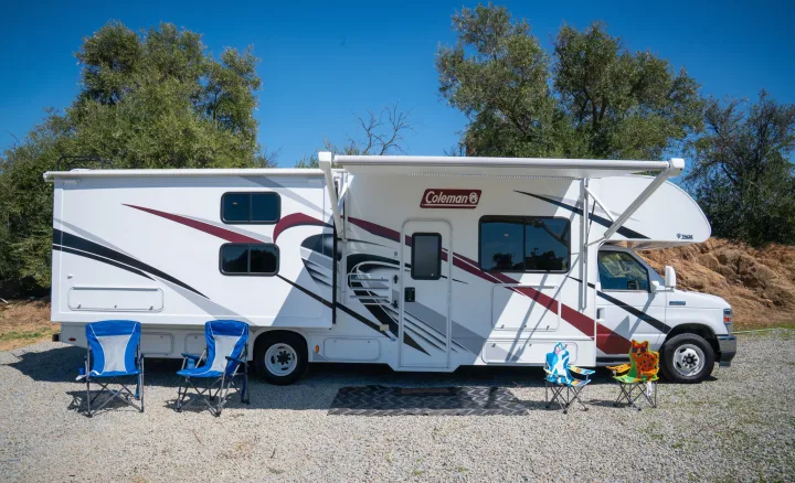 A white Coleman rv rental with an extended awning and camping chairs, parked on gravel, set for an outdoor leisure trip.. Thor Motor Coach Coleman 2023