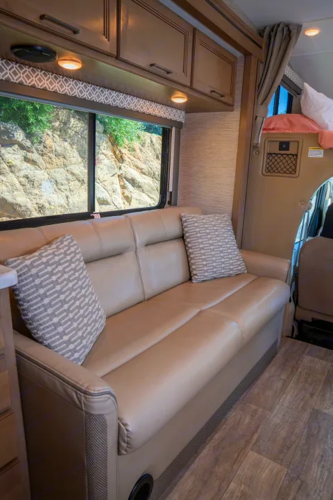 RV rental's inviting interior: beige sofa with patterned pillows, wood-look floor, and window revealing a rocky landscape.. Thor Motor Coach Coleman 2023