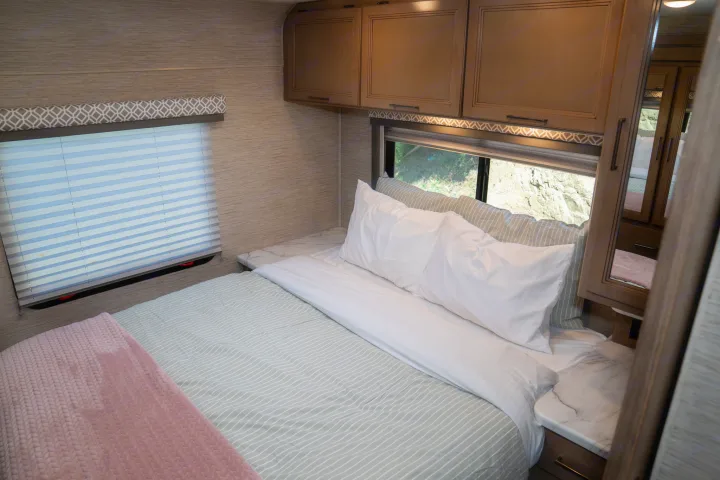 An RV rental's cozy bedroom features striped bedding, white pillows, overhead storage, and a window with an outdoor view.. Thor Motor Coach Coleman 2023