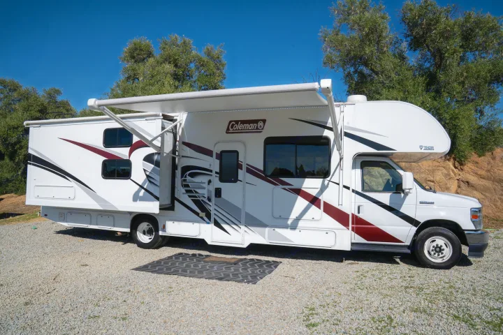 White Coleman rv rental (Thor Class C) with awning extended, parked on gravel under clear blue sky near trees.. Thor Motor Coach Coleman 2023