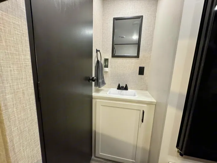 This RV rental features a compact bathroom with a white sink, modern black faucet, mirror, and dark entry door.. Jayco 260BH 2025
