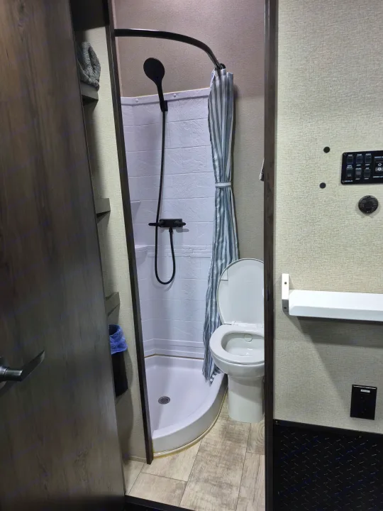 This RV rental bathroom features a compact shower, toilet, and modern control panel. Wood-look floor.. Grand Design Momentum 2022