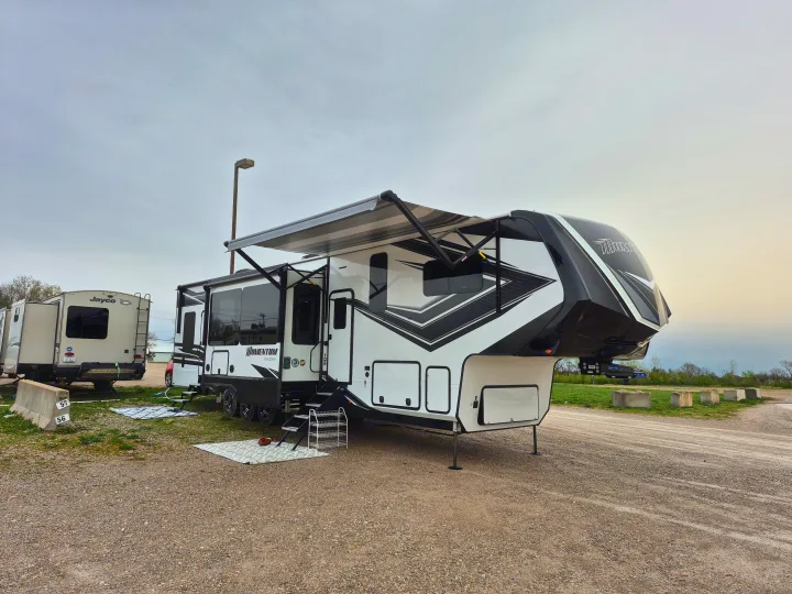 White & black Grand Design Momentum fifth-wheel RV parked at a gravel campsite with awning extended. Great rv rental!. Grand Design Momentum 2022