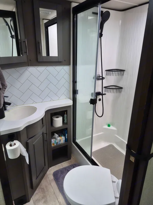 Compact RV rental bathroom with dark wood vanity, white sink, modern herringbone tile backsplash, toilet, and glass shower.. Grand Design Momentum 2022