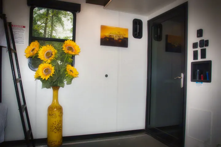 RV rental interior: sunflowers in a yellow vase, window with tree view, sunset painting, door, and wall controls. A bright, practical space.. Alliance Valor 2022
