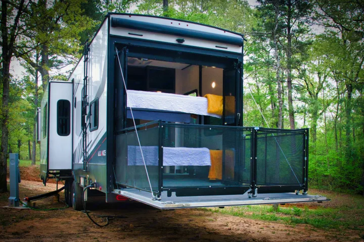 This toy hauler RV rental is parked in a forest with its rear ramp forming a patio, revealing bunk beds inside. Utility hookups are visible.. Alliance Valor 2022