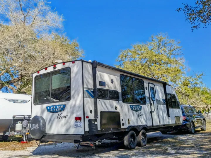 Sunny day scene: A large white travel trailer (RV rental) and attached SUV are parked amongst trees, ideal for camping.. Forest River Salem Cruise Lite 2024