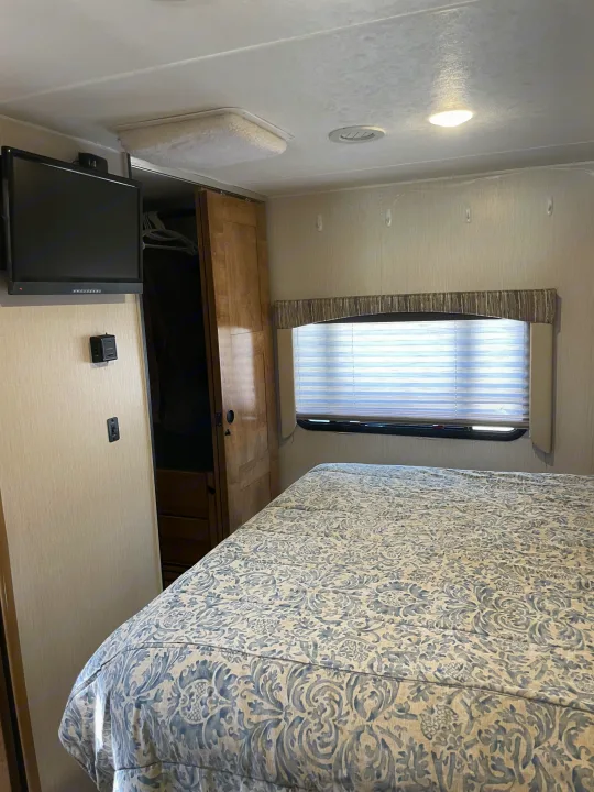This RV rental bedroom features a patterned bed, wall-mounted TV, and a window with blinds, showing a compact but functional space.. Forest River Coachmen Leprechaun 2020