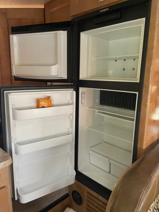 RV rental Dometic refrigerator with open doors, revealing empty freezer and fridge compartments, shelves, and a single box.. Coachmen Leprechaun 2021