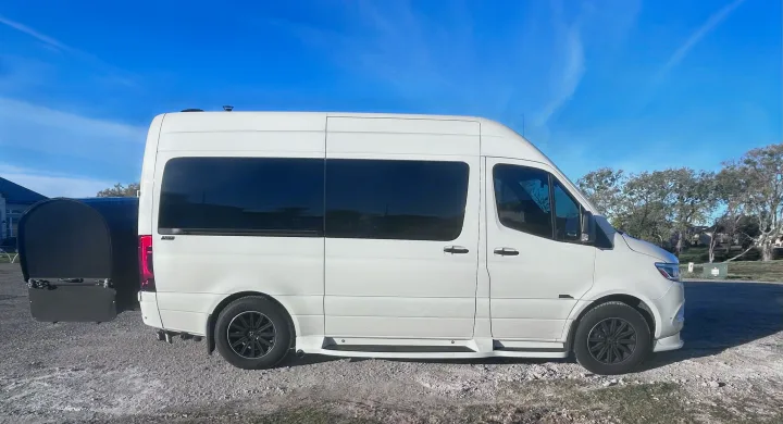 White Sprinter van, commonly used for RV rental, with a rear cargo carrier, parked on gravel under a blue sky.. Midwest Automotive Designs Luxe Cruiser 2024