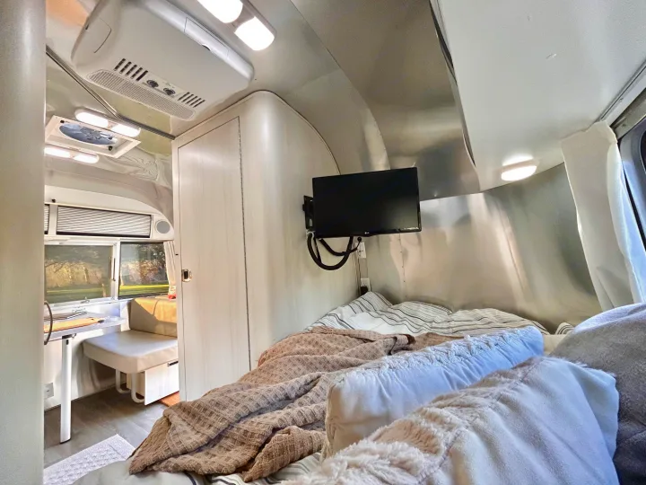 A bright, modern rv rental interior. The bedroom features a cozy bed with throws & a TV. A small dinette/kitchen area is visible through the doorway.. Airstream Sport 2018