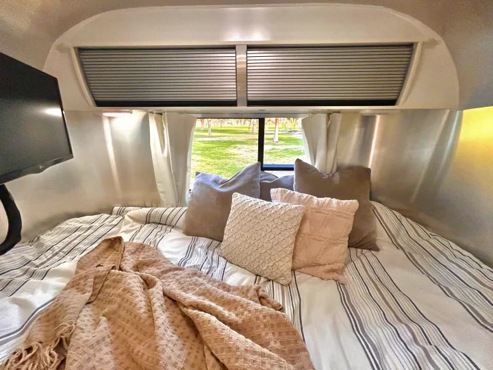 Cozy RV rental bed with striped bedding, various pillows, and a blanket, offering a relaxing view of nature.. Airstream Sport 2018