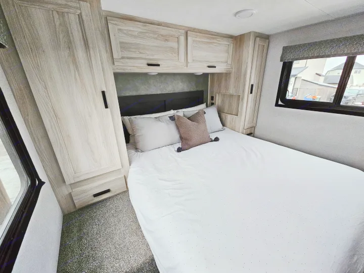 Modern RV rental bedroom with a large bed, light wood cabinets overhead and flanking it, and a window to the right. Grey carpet.. Forester 2651 CDW 2022