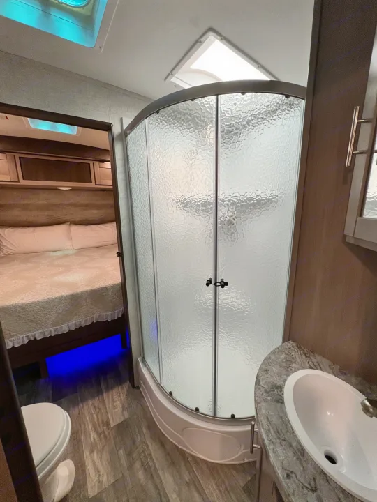Interior of an RV rental showing a full bathroom with a curved shower, sink, and toilet, plus a bedroom with blue-lit under-bed area.. Crossroads RV Sunset Trail Super Lite 2021