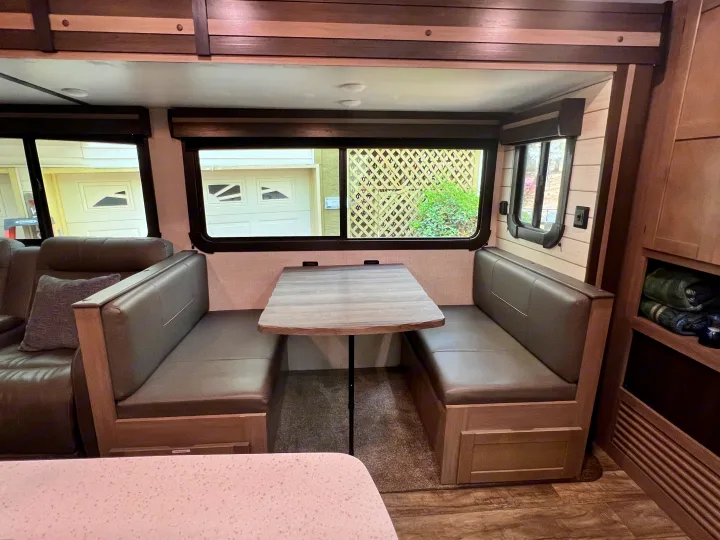 This RV rental features a cozy dinette with grey upholstered benches and a wooden table, ideal for dining or relaxing by the windows.. Crossroads RV Sunset Trail Super Lite 2021