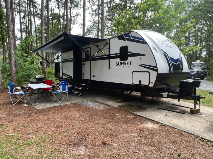 A white travel trailer RV rental, with an extended awning, parked at a wooded campsite with a picnic area.. Crossroads RV Sunset Trail Super Lite 2021