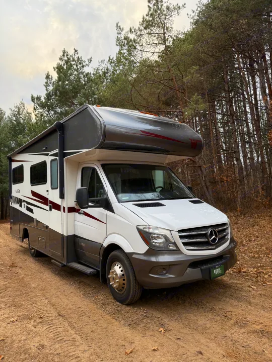 A white and gray Winnebago Vita RV rental is parked on a dirt road, surrounded by tall pine trees under an overcast sky.. Winnebago Vita 2020