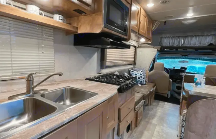 Modern RV rental interior: kitchen with double sink, gas stove, microwave, comfortable dinette, and front cab view.. Coachmen Leprechaun 260RS 2020