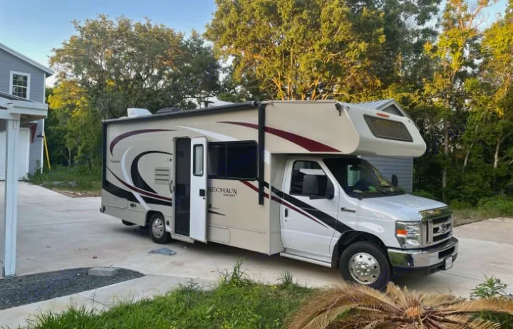 A beige & white Class C motorhome, an excellent rv rental option, parked on a driveway. Open door, lush trees, and house visible.. Coachmen Leprechaun 260RS 2020