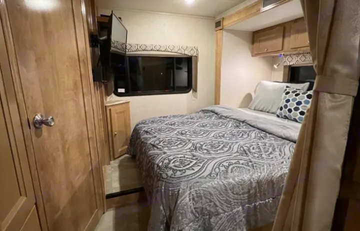 Cozy RV rental bedroom features a patterned bed, wall-mounted TV, window, and wood cabinetry, ideal for compact travel.. Coachmen Leprechaun 260RS 2020