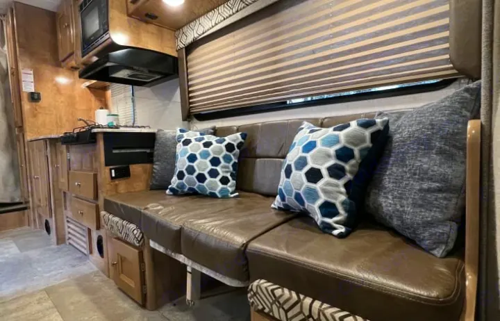 This RV rental features a compact kitchen with a stove and microwave, and comfortable seating with blue/gray hexagonal pillows.. Coachmen Leprechaun 260RS 2020