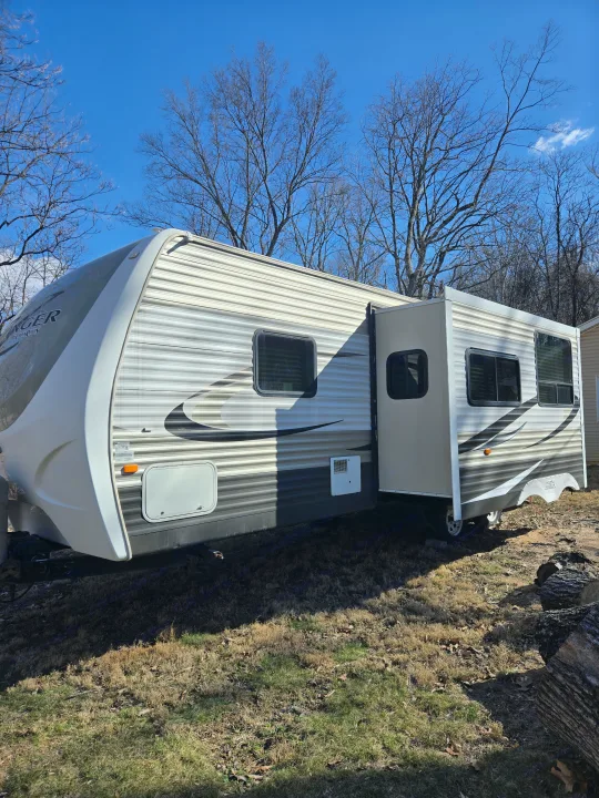 A modern, white & gray travel trailer RV with an extended slide-out, parked on grass beneath bare trees & blue sky. Consider for rv rental.. Crossroads RV Zinger 2014