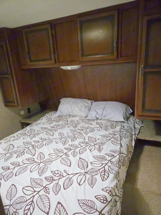 Inside an rv rental: a cozy bedroom with leaf-patterned bedding, two pillows, and dark wood cabinetry overhead.. Crossroads RV Zinger 2014