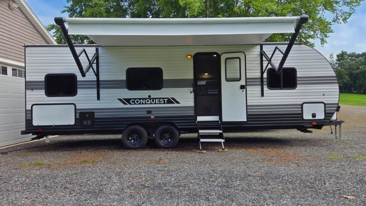 Spacious RV rental, a Conquest travel trailer, with its awning extended and door open, ready for adventure on a gravel lot.. Gulf Stream Trailmaster 2024
