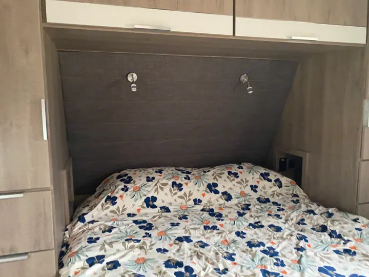 View of a cozy RV rental bed with a blue and white floral duvet, surrounded by built-in storage and reading lamps.. Grand Design Imagine 2021