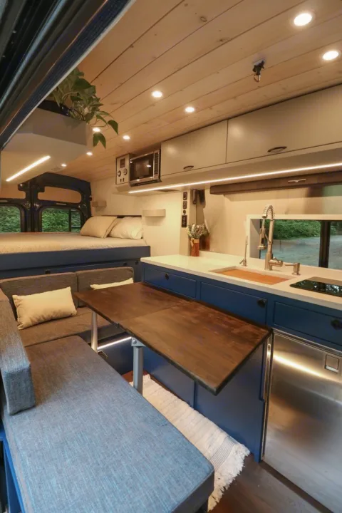 This modern RV rental interior features a cozy bed, dining area with a folding table, and a compact kitchen.. Ford Transit 2016