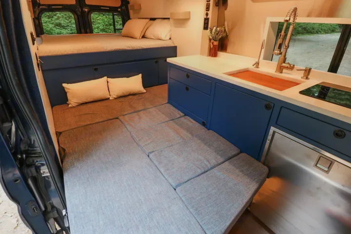 Modern RV rental interior showing a comfortable bed, convertible seating area, and a fully equipped kitchen with a sink & fridge.. Ford Transit 2016