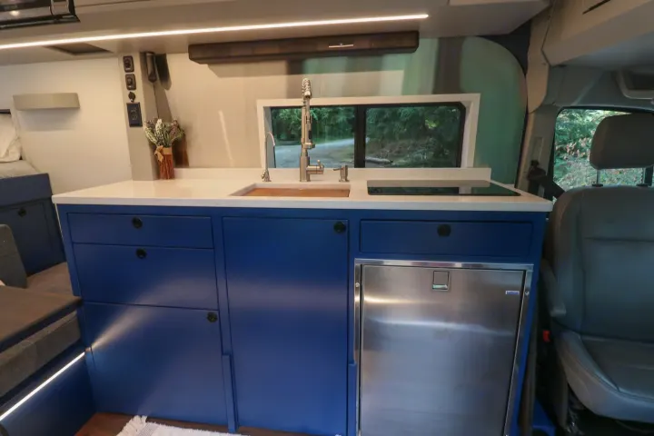 Inside an RV rental: a modern kitchen with blue cabinets, white counters, sink, cooktop, and a view of nature.. Ford Transit 2016