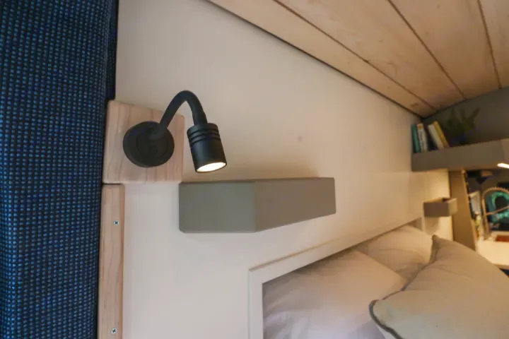 Close-up of an RV rental bedroom showing a black reading lamp, light wood accents, bed, and gray floating shelf.. Ford Transit 2016
