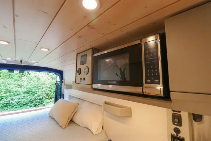This RV rental interior features a Black+Decker microwave and control panel above a cozy bed, with a view of lush greenery.. Ford Transit 2016
