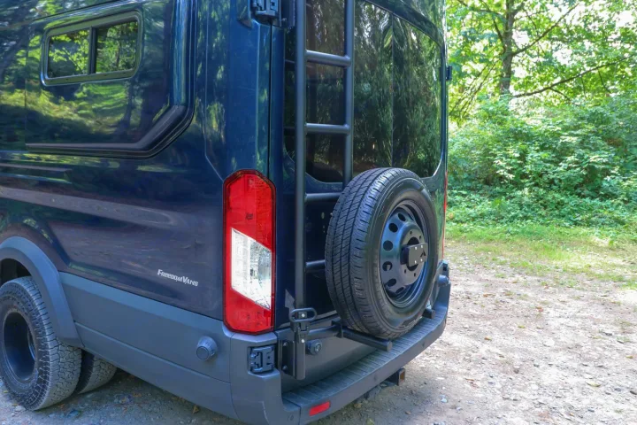 Rear view of a dark blue FreedomVANS camper, featuring a ladder and rear-mounted spare tire, perfect for RV rental.. Ford Transit 2016