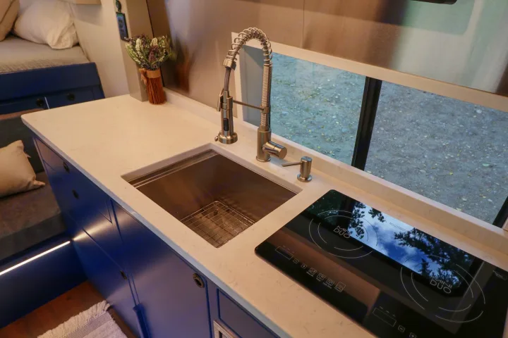 Modern RV rental kitchen featuring sleek white counters, a sink, and induction cooktop. Blue cabinets brighten the space.. Ford Transit 2016
