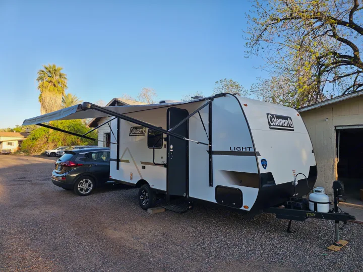 A bright white Coleman travel trailer with awning extended, parked next to a dark grey hatchback car, ready for RV rental.. Keystone Coleman 17BX 2025