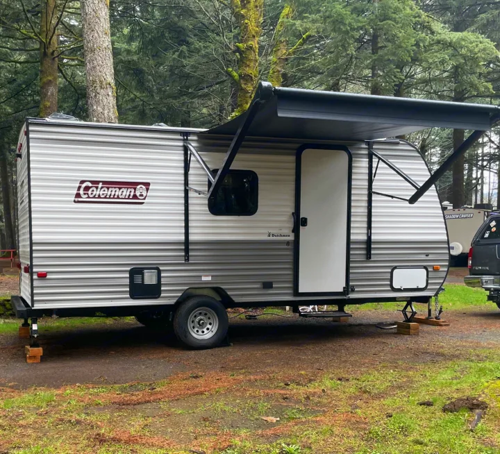 A Coleman RV rental travel trailer with its awning extended, stabilized at a green, wooded campsite.. Coleman 17B 2024