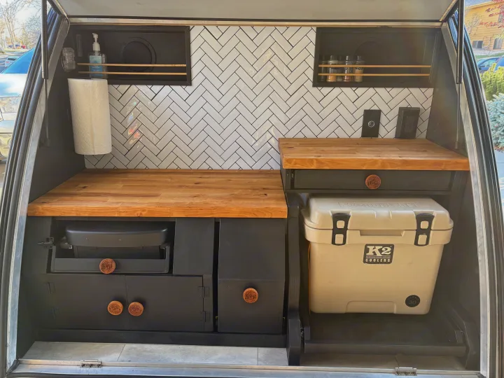 A compact, stylish RV rental kitchen featuring wood countertops, herringbone tiles, and built-in cooking gear and cooler.. Braxton Creek Free Solo 2022