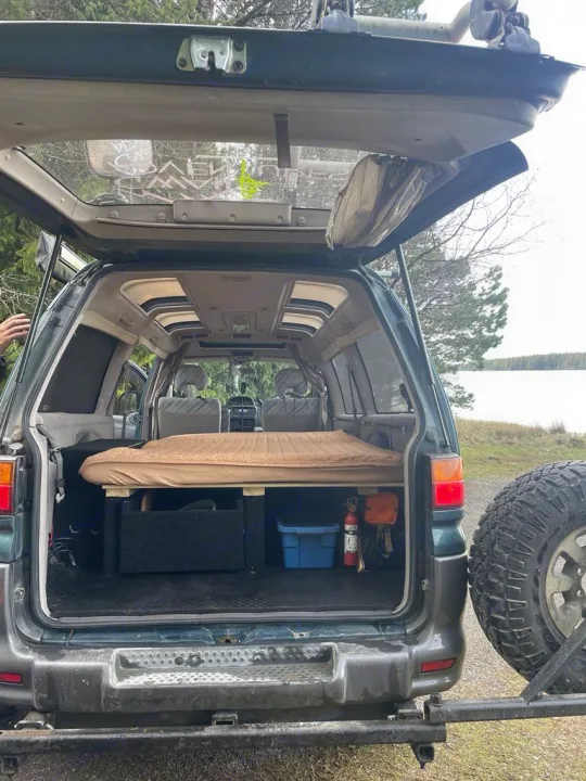 Open tailgate of a dark green overland van RV rental, showing a bed platform, storage, and safety gear for adventure trips.. MITSUBISHI Delica 1997