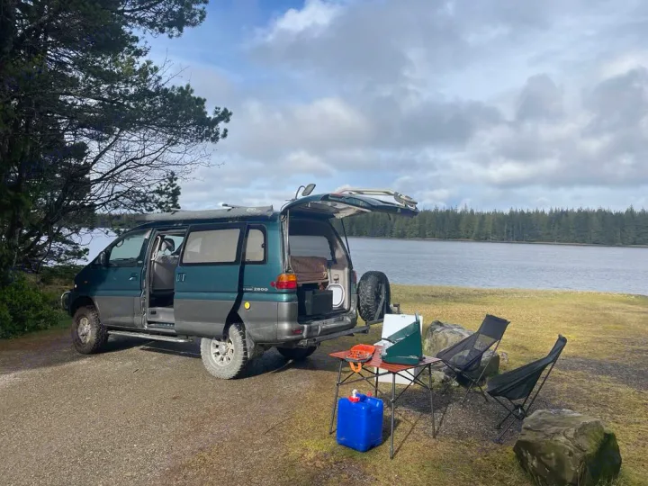 Dark teal RV rental camper van (Mitsubishi Delica) by a serene lake with camping gear setup for outdoor adventure.. MITSUBISHI Delica 1997