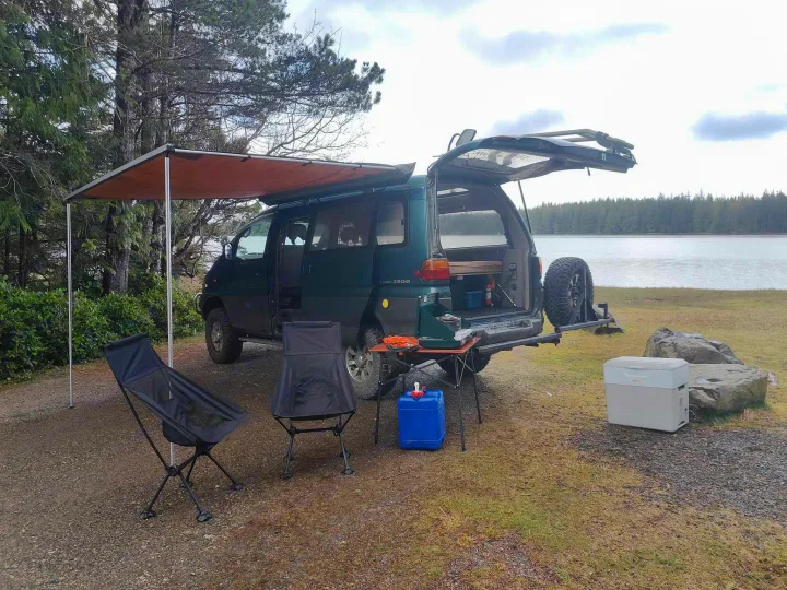 RV rental camper van with awning set up by a serene lake. Camping chairs, stove, and cooler ready for outdoor adventure!. MITSUBISHI Delica 1997