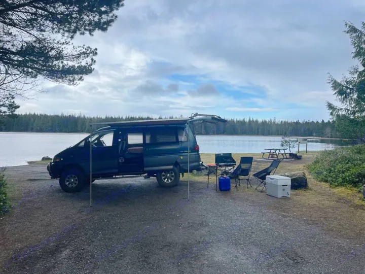 A dark camper van with an awning extended, parked by a lake at a campsite with chairs and gear. Great RV rental idea!. MITSUBISHI Delica 1997