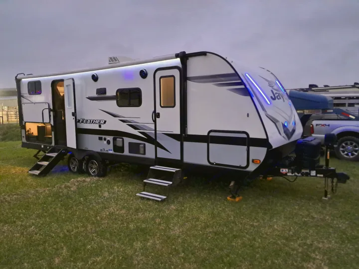 A modern white travel trailer, an ideal rv rental, illuminates the grass at dusk with blue and warm interior lights.. Jayco Jay Feather 2022
