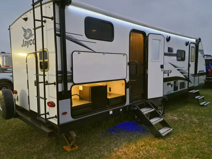 Brightly lit RV rental at dusk, showcasing its outdoor kitchen compartment, awning LEDs, and illuminated steps.. Jayco Jay Feather 2022