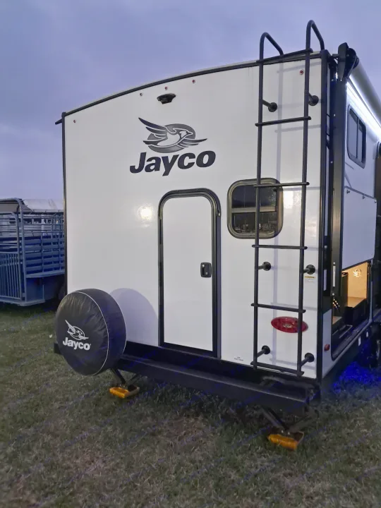 Rear view of a white Jayco travel trailer RV rental, showing its ladder, spare tire, and entry door.. Jayco Jay Feather 2022