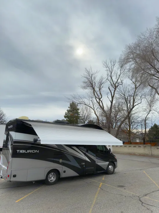 A black and silver RV rental with its awning extended is parked under an overcast sky with bare trees.. Thor Tiburon 2022