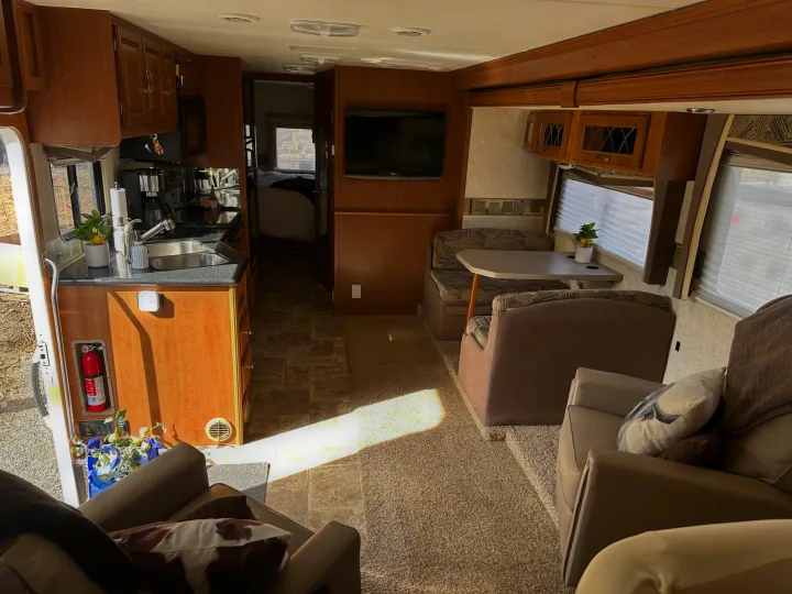 Bright RV rental interior. Features kitchen, dining booth, comfy living space, and a visible bedroom. Wood cabinetry and sunlit carpet.. Forest River Georgetown 2011