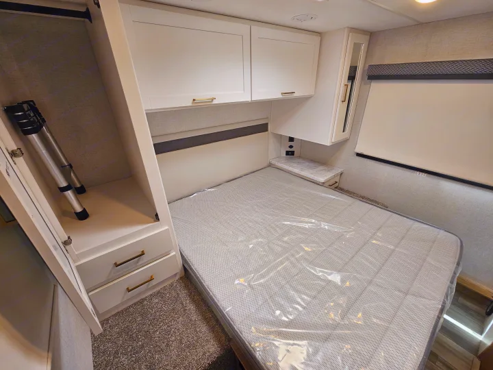 Modern RV rental bedroom: a bed with plastic cover, white storage cabinets, nightstand, and wardrobe with a tripod inside.. Thor Motor Coach Four Winds 2026